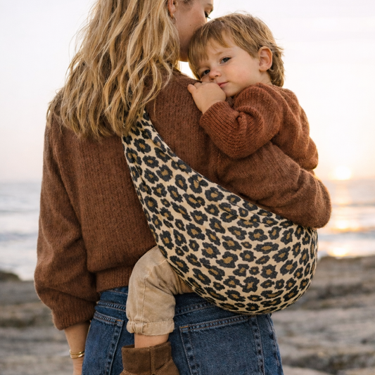 mama blonde qui porte son bébé dans un sling vu de dos à la page. Le bébé est apaisé en toute serenité