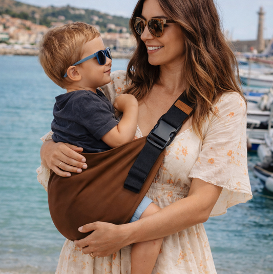 maman brune tient son bébé dans un porte-bébé sling au bord de la plage. Son enfant est souriant (la position de son dos et son bassin est respectée)