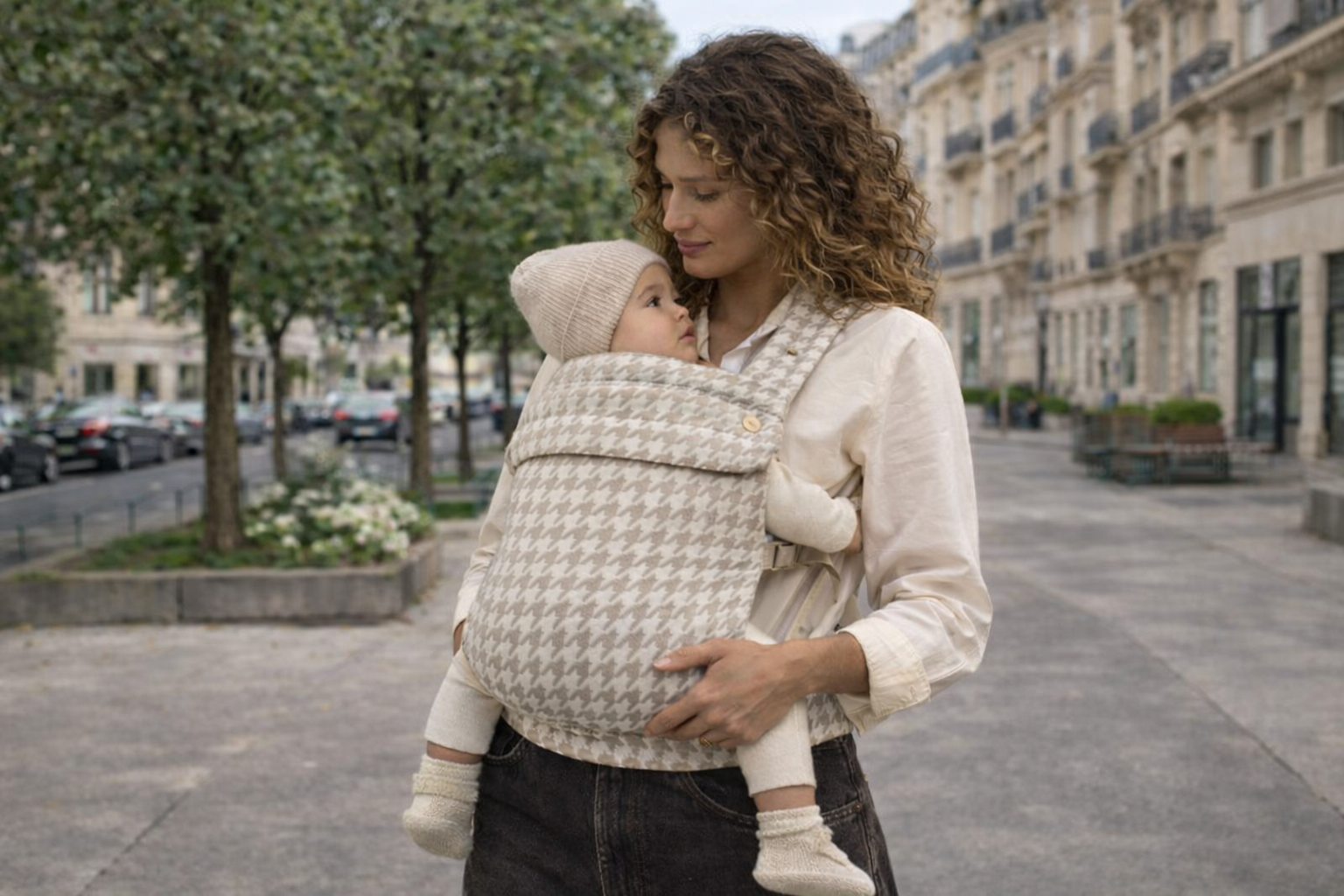 maman bouclée qui tient son bébé dans un porte bébé physiologique au motif pied-de-poule coloris beige