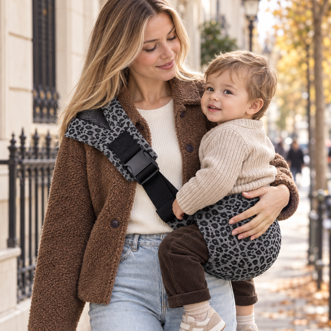 Maman blonde avec une veste moumoute qui tient son enfant dans un sling léopard gris