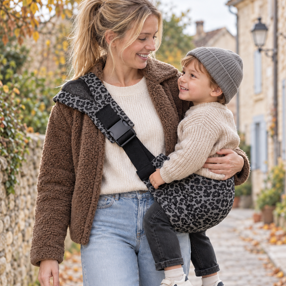 Maman blonde avec une veste moumoute qui tient son enfant dans un sling léopard gris