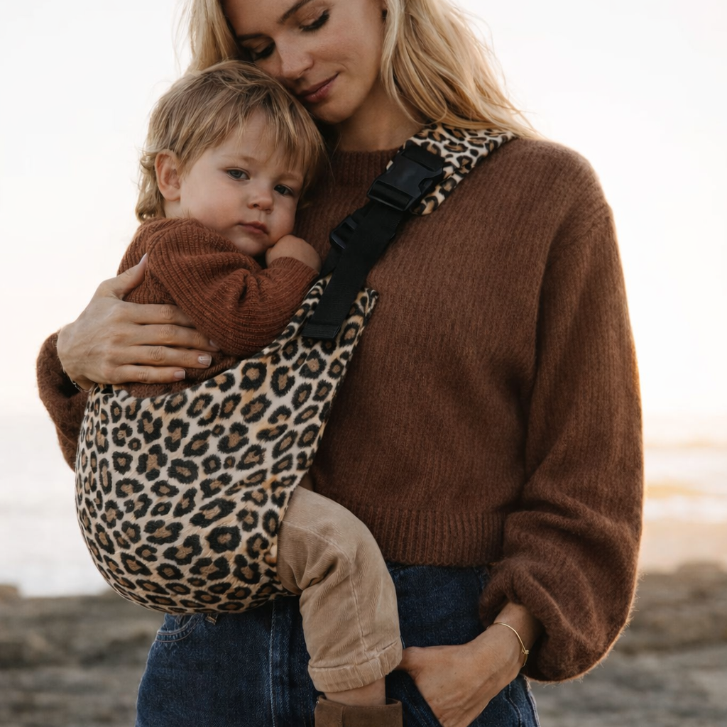 Maman blonde porte son bébé dans un porte bébé sling couleur léopard beige à la mer