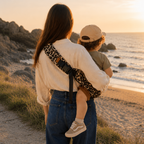 Maman brune porte son bébé dans un porte bébé sling couleur léopard beige à la mer sur une plage bretonne, vue de dos