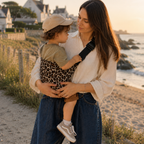 Maman brune porte son bébé dans un porte bébé sling léopard beige à la mer sur une plage bretonne
