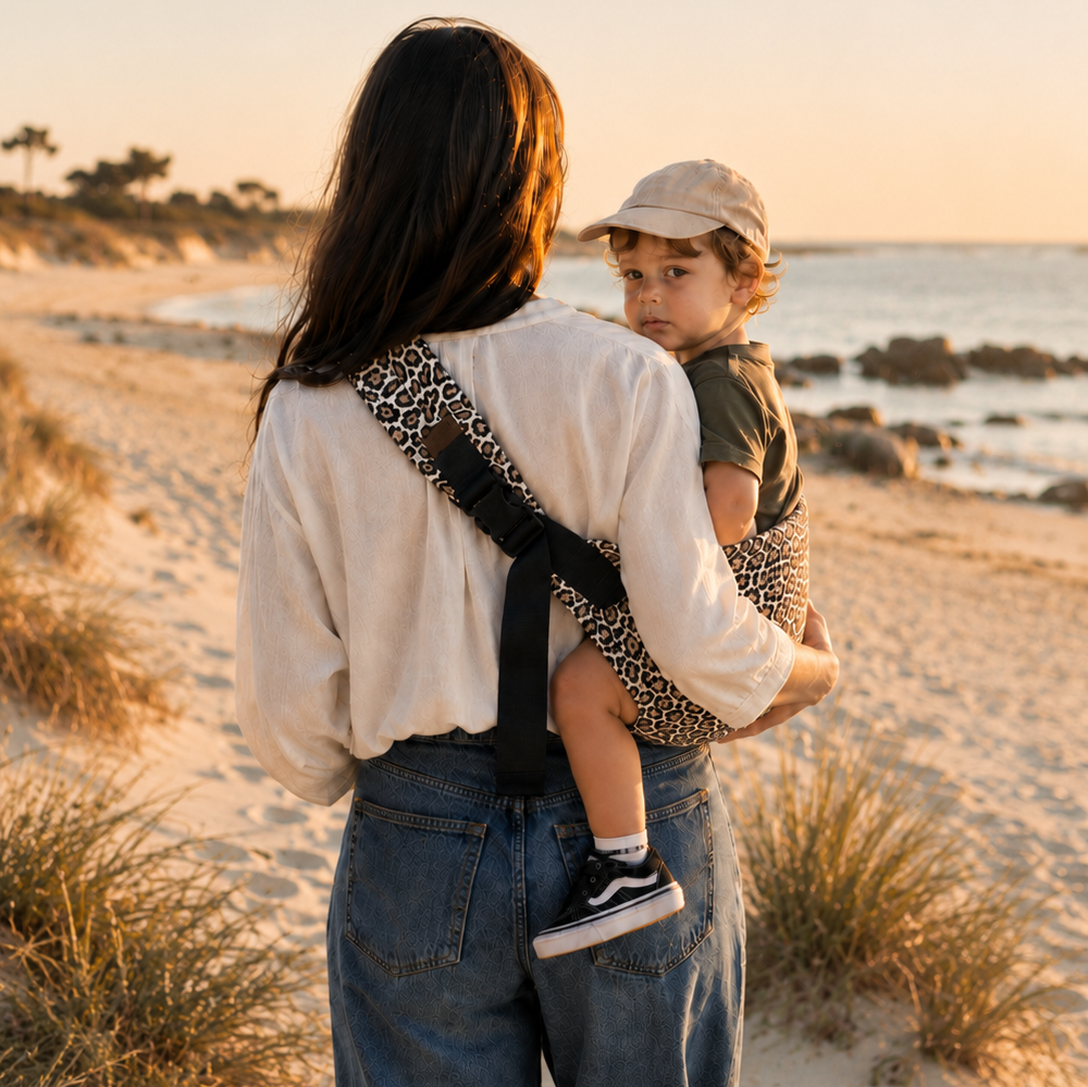 Maman brune porte son bébé dans un porte bébé sling couleur léopard beige à la mer sur une plage bretonne, vue de dos