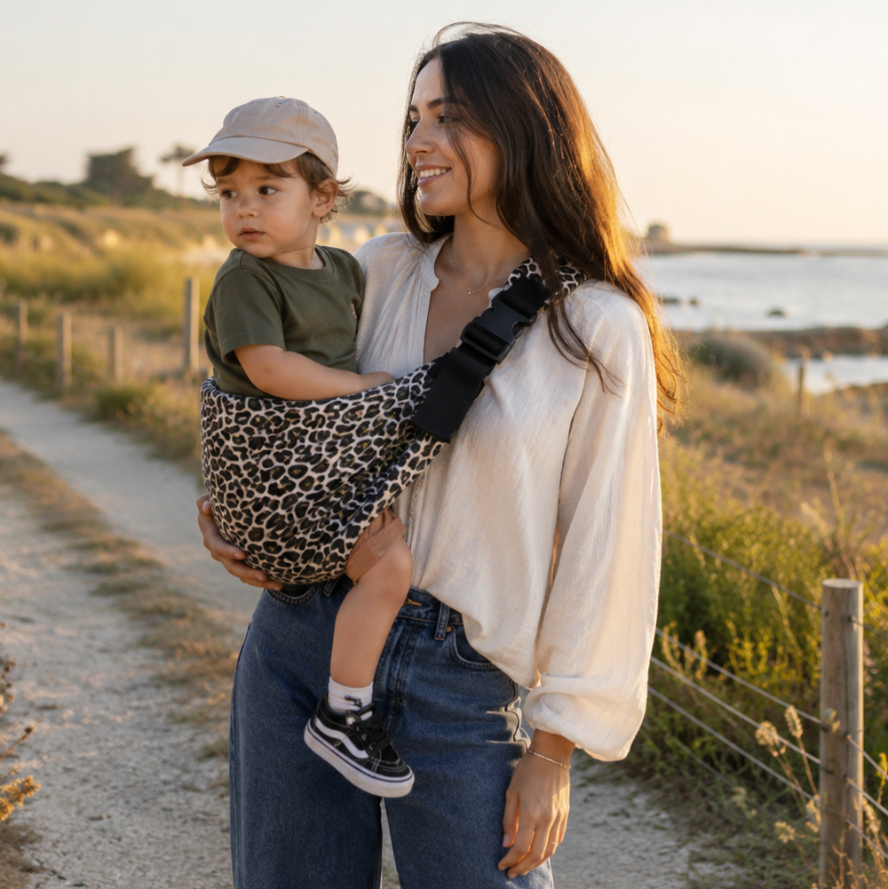 Maman brune porte son bébé dans un porte bébé sling léopard beige à la mer sur une plage bretonne