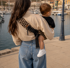 Maman brune porte son bébé dans un porte bébé sling couleur léopard beige devant un port, vue de dos 