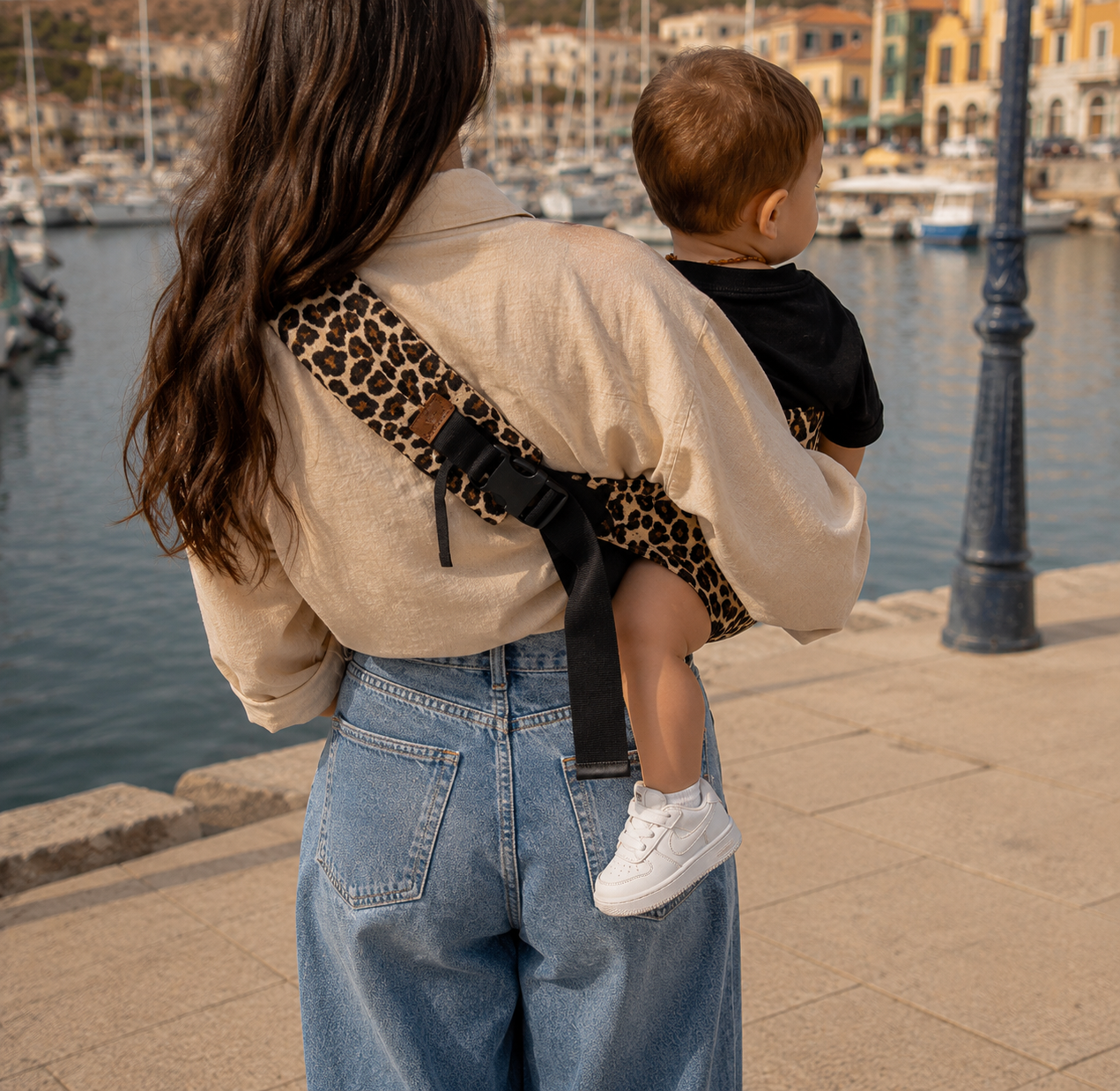 Maman brune porte son bébé dans un porte bébé sling couleur léopard beige devant un port, vue de dos 