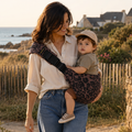 Maman brune qui se balade sur la plage avec son bébé dans les bras. Il est dans un porte-bébé sling léopard moka, apaisé et souriant.