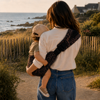 Maman brune qui se balade sur la plage avec son bébé dans les bras. Il est dans un porte-bébé sling léopard moka, apaisé et souriant. Vue de dos pour voir le sling