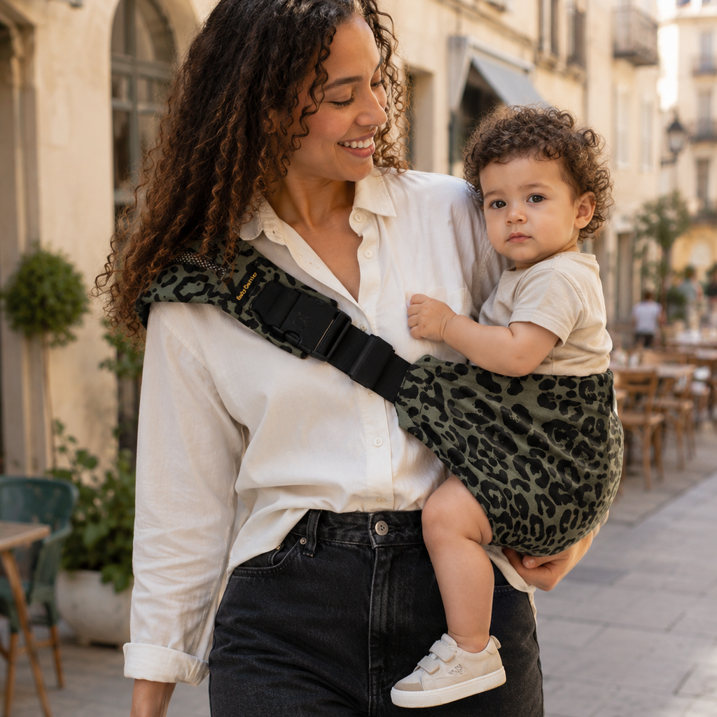 Maman métisse se promène avec son enfant aux cheveux bouclés. Elle le porte dans un porte-bébé sling de couleur léopard olive. Il était fatigué et maintenant apaisé