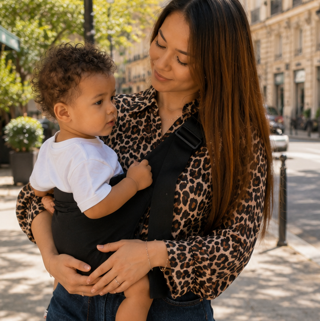 Femme métisse avec chemise léopard tient son bébé avec un porte bébé noir, dans la rue au printemps