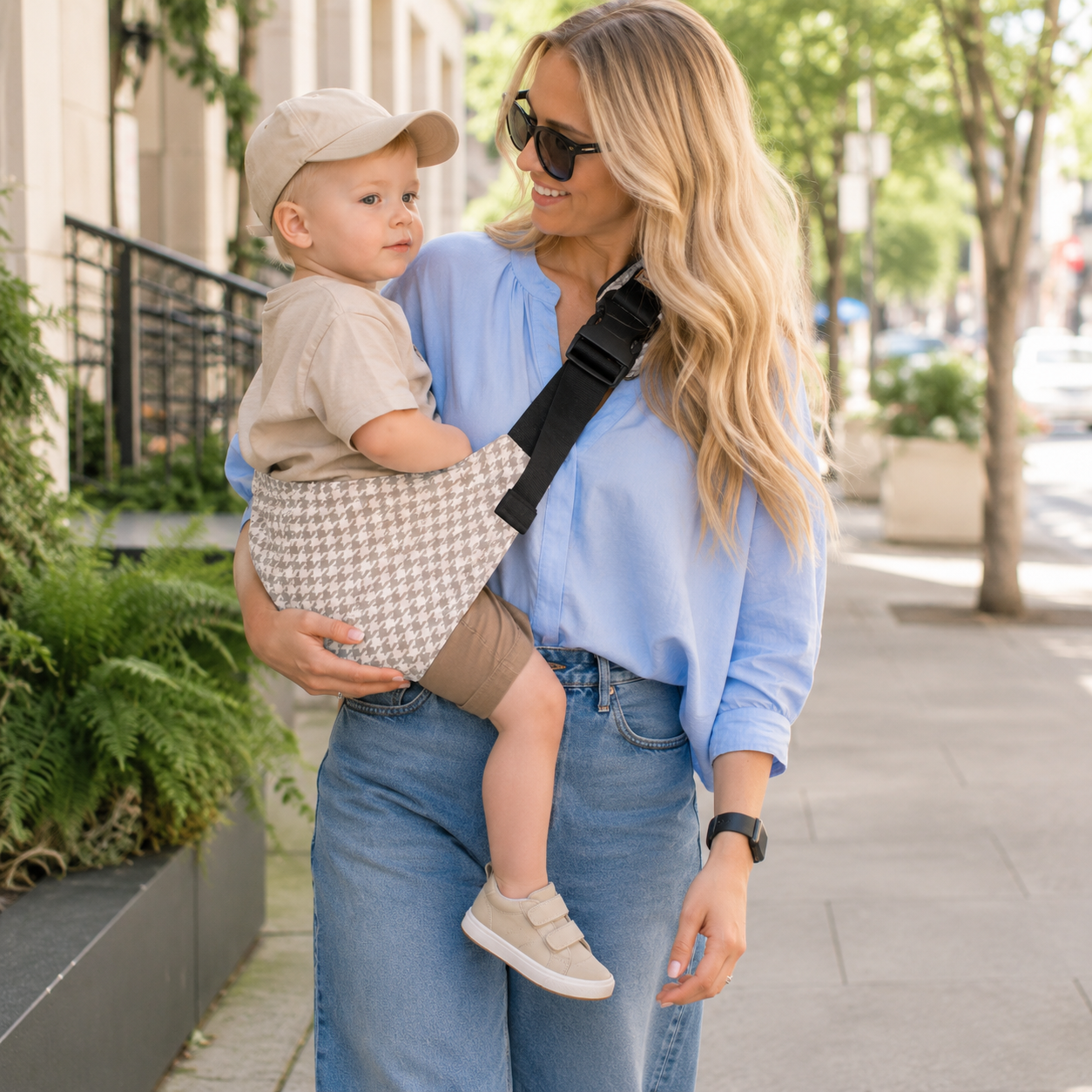 Maman blonde qui tient son petit garçon dans son porte-bébé sling motifs pied-de-poule beige durant l'été en pleine ville