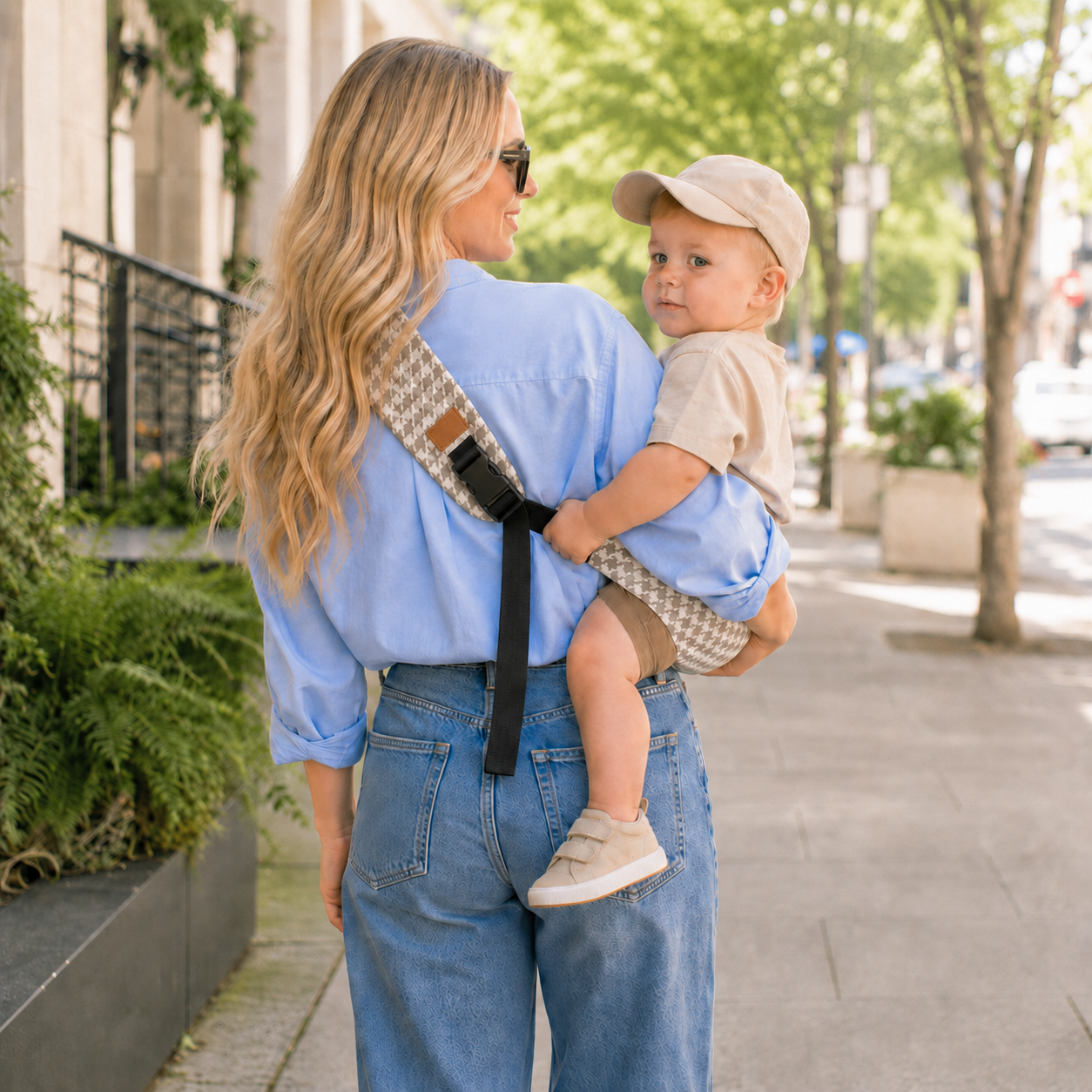 Maman blonde qui tient son petit garçon dans son porte-bébé sling motifs pied-de-poule beige durant l'été en pleine ville, vue de dos