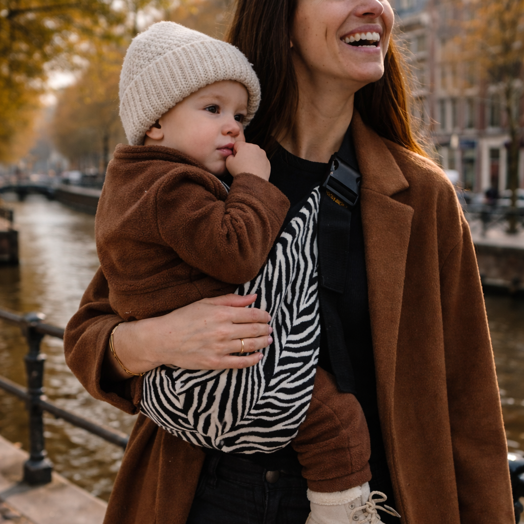 Maman brune qui tient son bébé dans un porte-bébé zebre. Le bébé porte un bonnet. Ils sont en ville durant l'automne