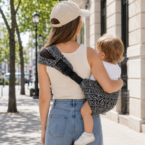 Maman portant une casquette balade son bébé dans la rue en été. Elle le porte dans un porte-bébé sling motifs aztek. Vue de dos pour le voir le sling