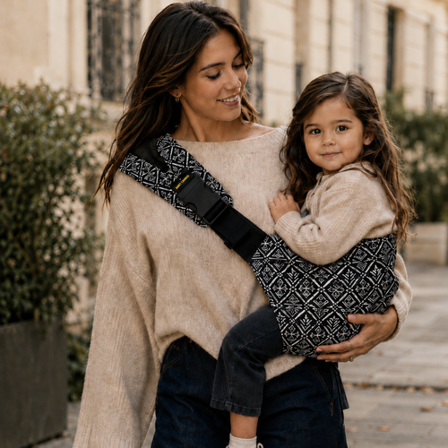 maman brune qui porte sa petite fille de 3 ans prête pour la rentrée. Elle est bien reposée dans le porte-bébé sling aztek
