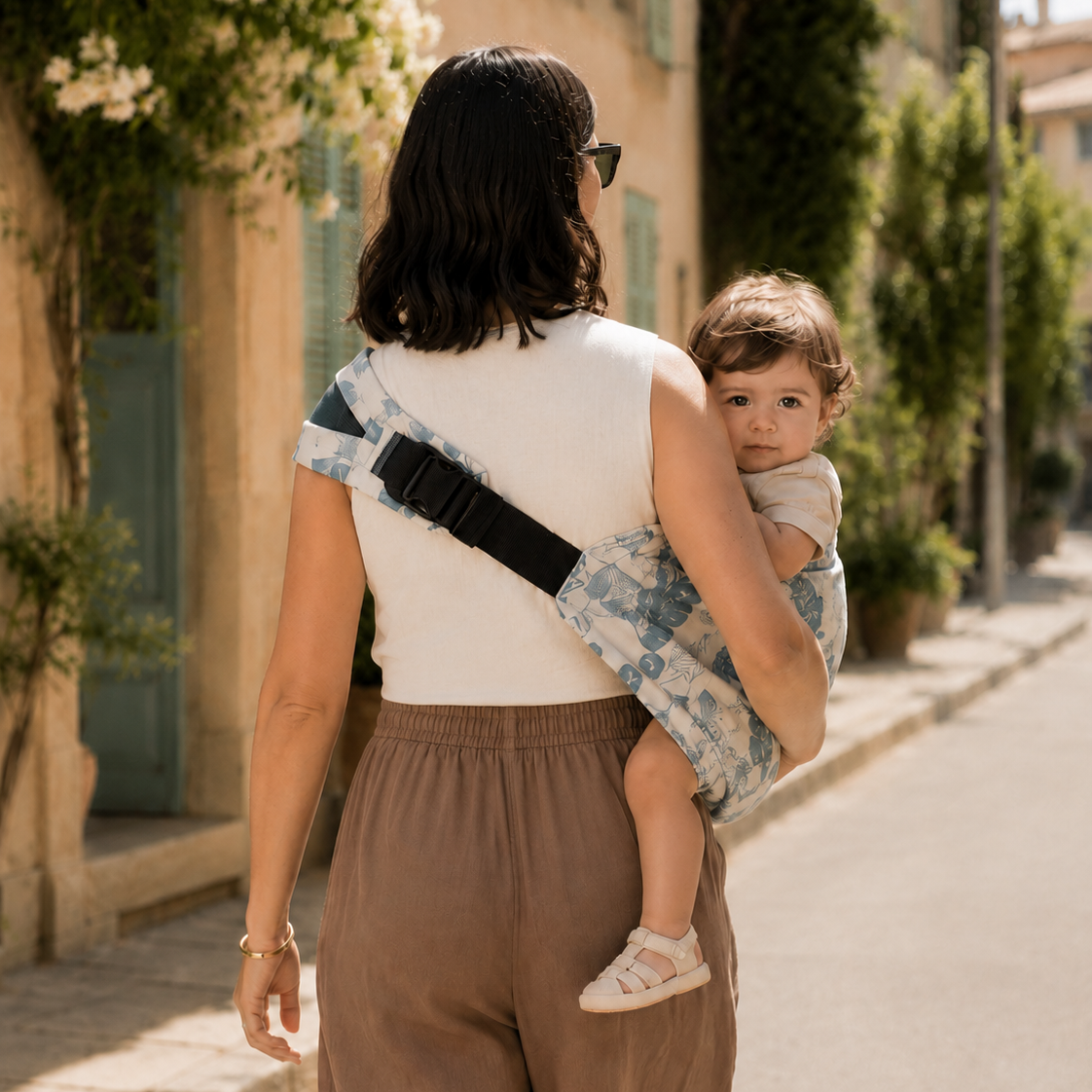 Maman aux cheveux noires porte son bébé chatain dans un porte-bébé sling style palmier. Ils marchent dans la rue sous le soleil. Vue de dos