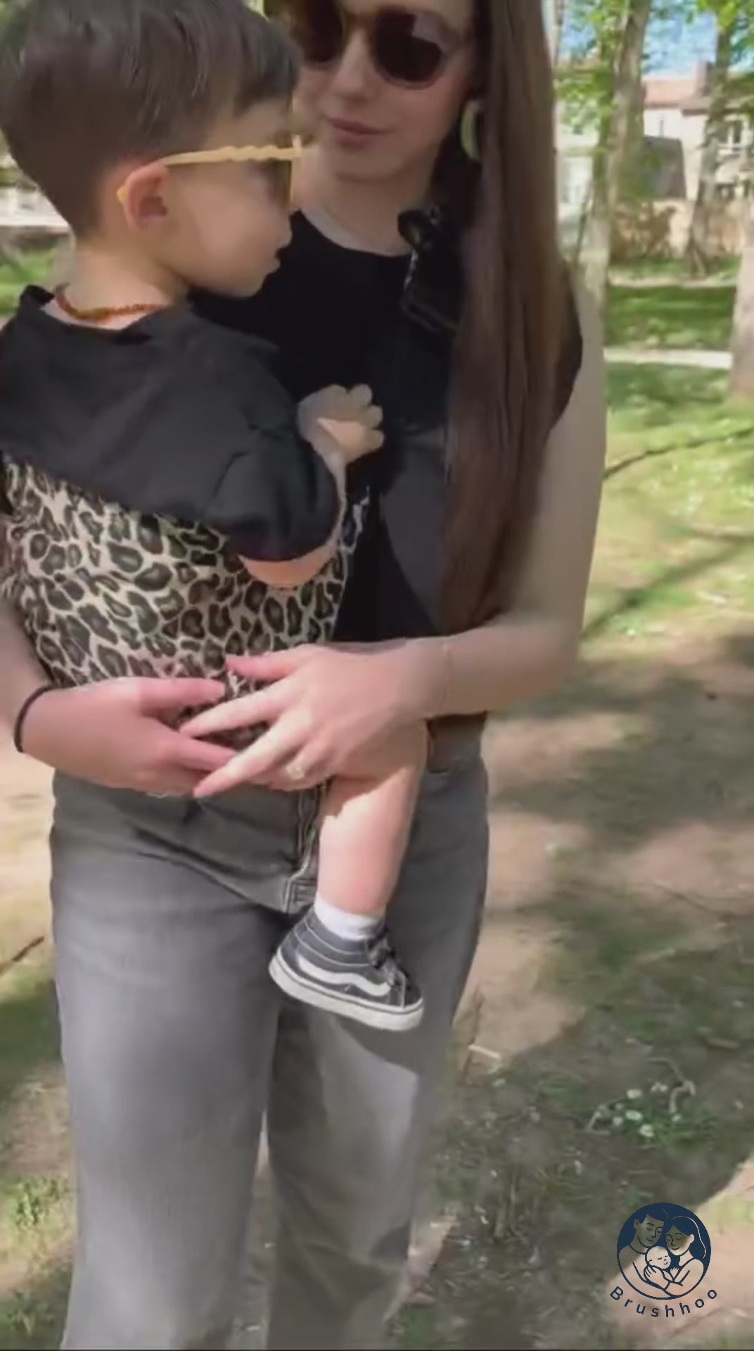 Maman brune qui porte son bébé dans un porte-bébé sling léopard ecru. Elle se balade l'esprit léger dans un parc